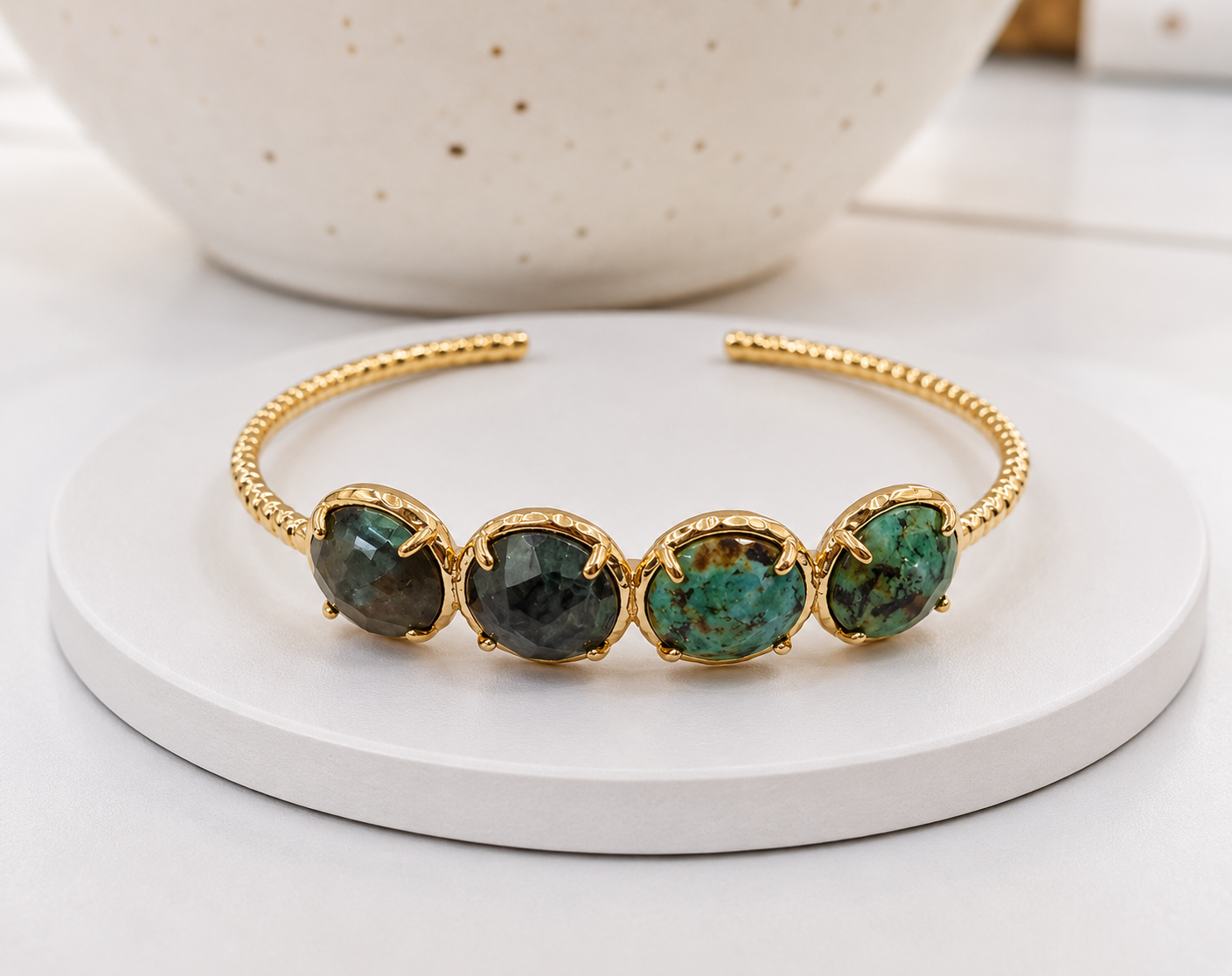 
                  
                    A gold-tone open cuff bracelet featuring four oval African Turqouise stones, displayed on a white round tray with a ceramic bowl in the background.
                  
                
