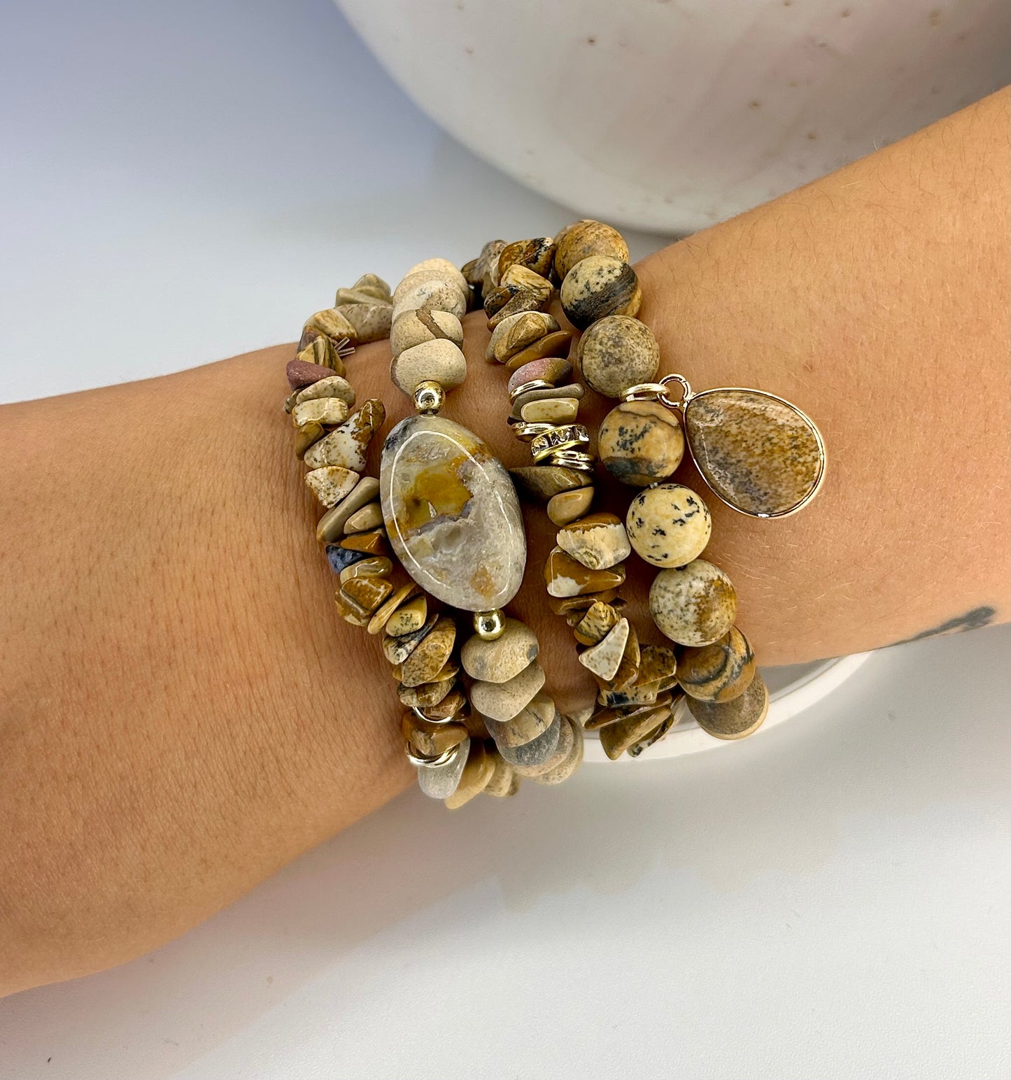 
                  
                    Stack of desert jasper stone and gold beaded bracelets on a wrist with a neutral background
                  
                