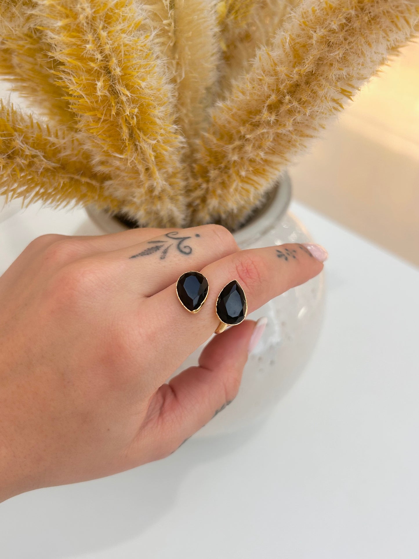 
                  
                    A close-up of a hand wearing a gold  adjustable ring with black obsidian teardrop-shaped stones, posed in front of a vase of dried pampas grass on a white surface.
                  
                