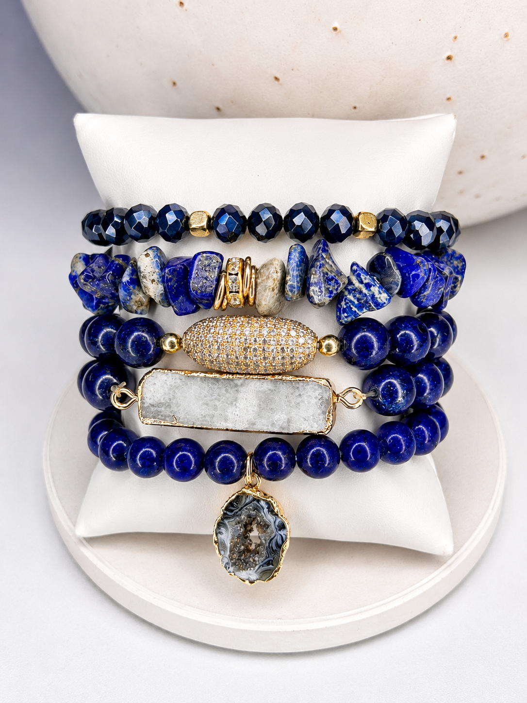 
                  
                    Stack of blue lapis lazuli beaded bracelets with gold accents on a white surface
                  
                