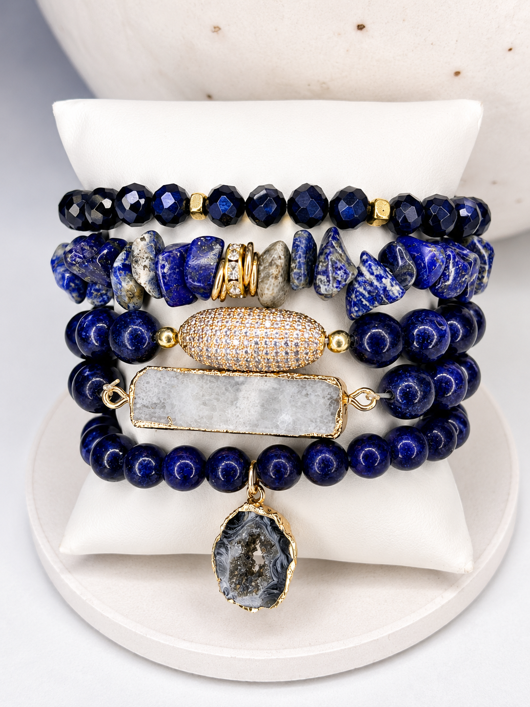 
                  
                    Stack of blue lapis lazuli mixed natural stone beaded bracelets with gold accents on a white surface
                  
                