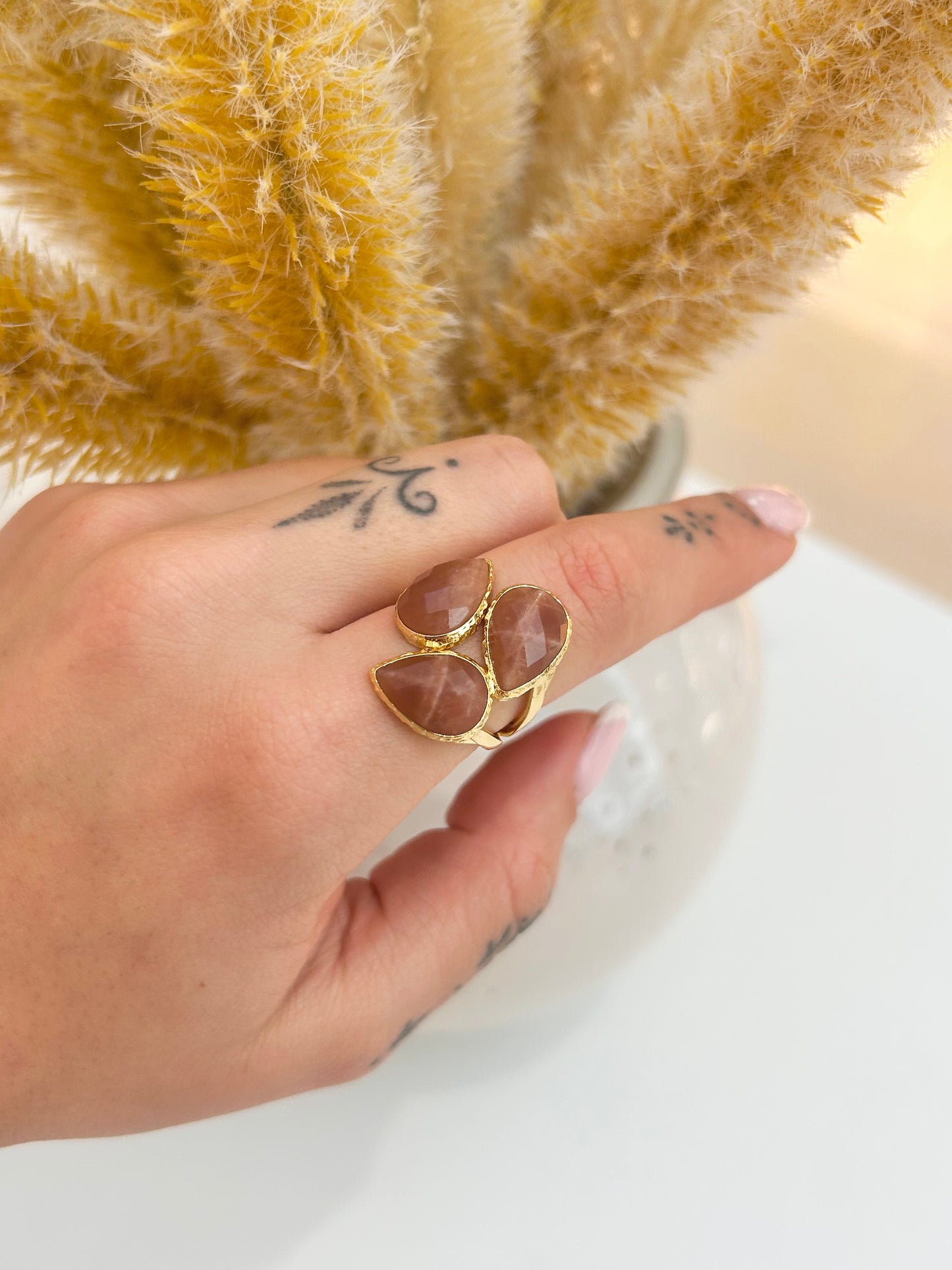 
                  
                    A close-up of a hand wearing a gold ring with three teardrop-shaped sunstone gemstones, set against a background of dried yellow pampas grass in a vase.
                  
                