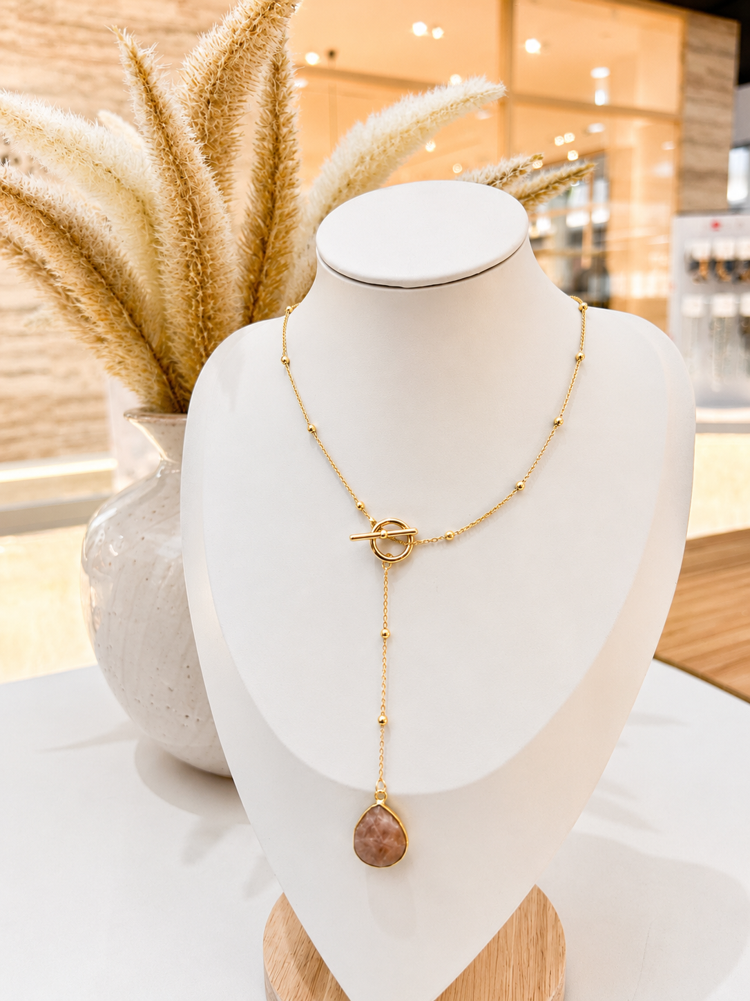 
                  
                    Gold necklace with a sunstone pendant on a mannequin bust in a store setting
                  
                