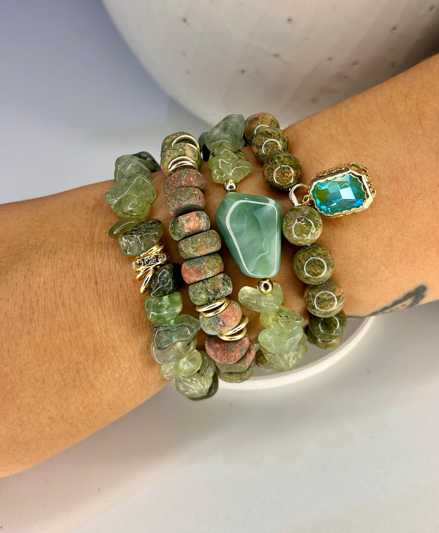 
                  
                    Stack of green stone beaded bracelets on a wrist with a neutral background
                  
                