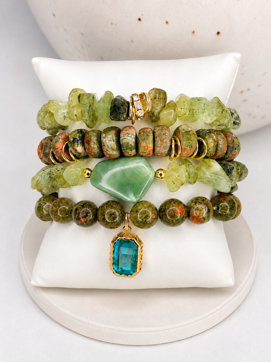 
                  
                    Four beaded stretch bracelets displayed on a white cushion. The bracelets feature natural green and brown jasper beads, a light green stone, and a central turquoise-blue rectangular gemstone pendant with gold-tone metal accents.
                  
                