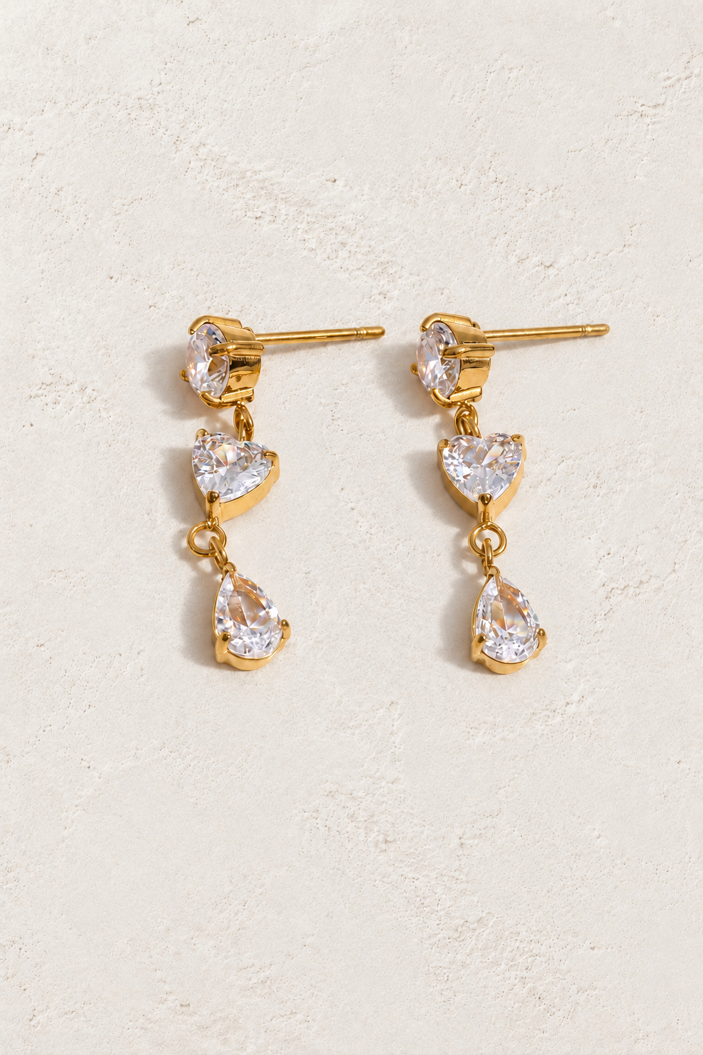 
                  
                    Gold three-stone heart and teardrop earrings on approved cream hero background
                  
                