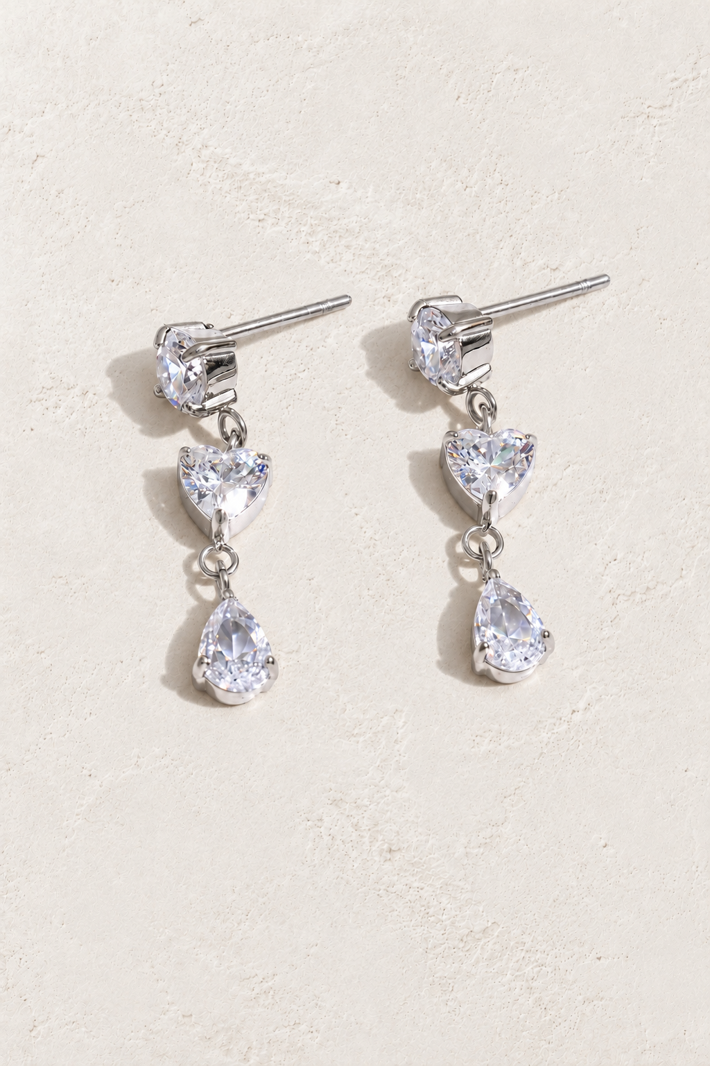 
                  
                    Silver three-stone heart and teardrop earrings on approved cream hero background
                  
                