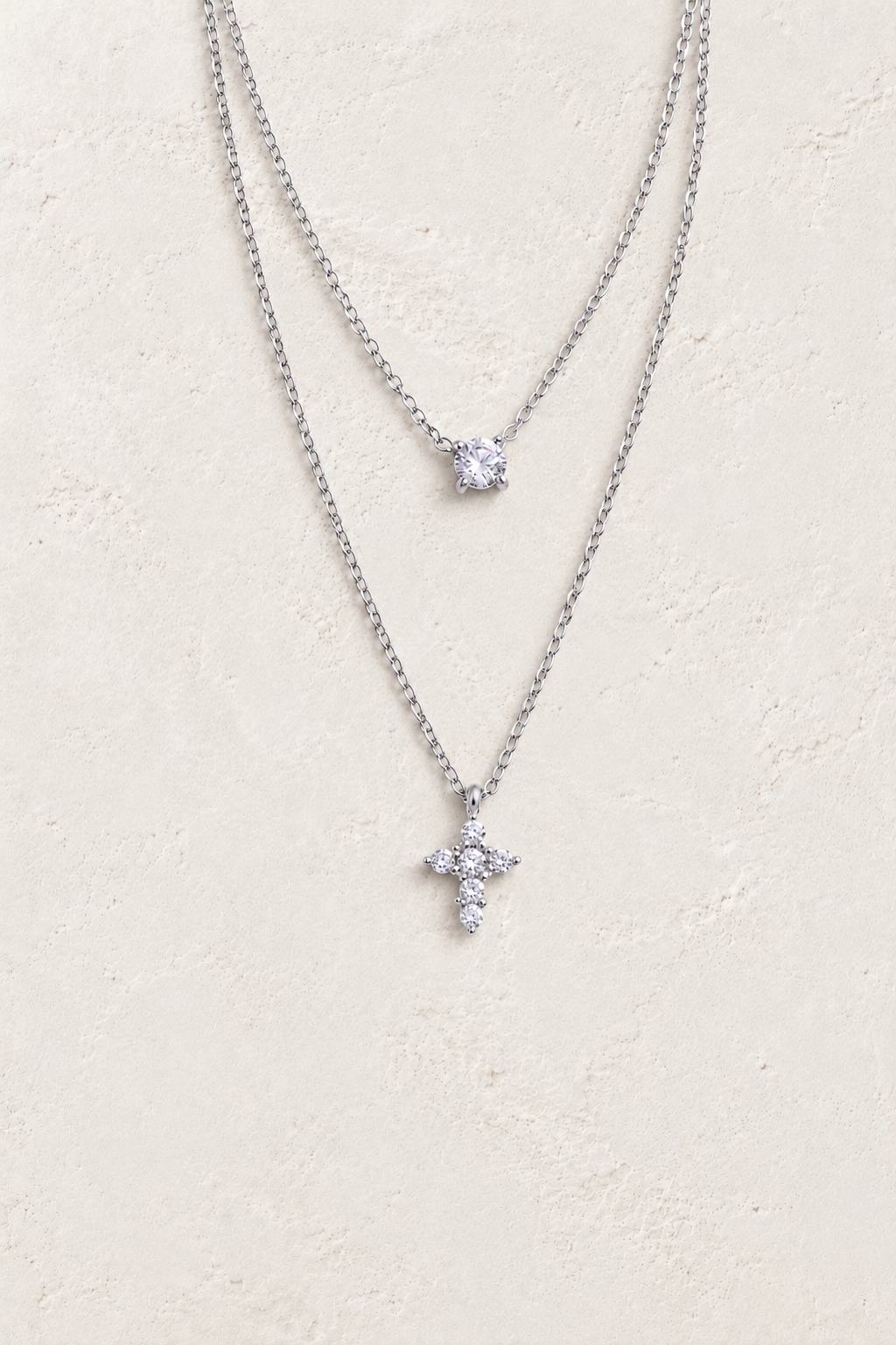 
                  
                    Silver layered solitaire and cross necklace on approved cream hero background
                  
                
