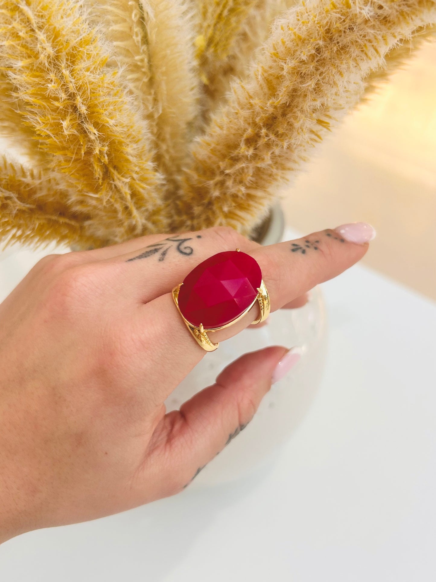 
                  
                    A gold ring with a large oval red gemstone worn on a person's finger, with a blurred background of dried beige grass and a white surface.
                  
                