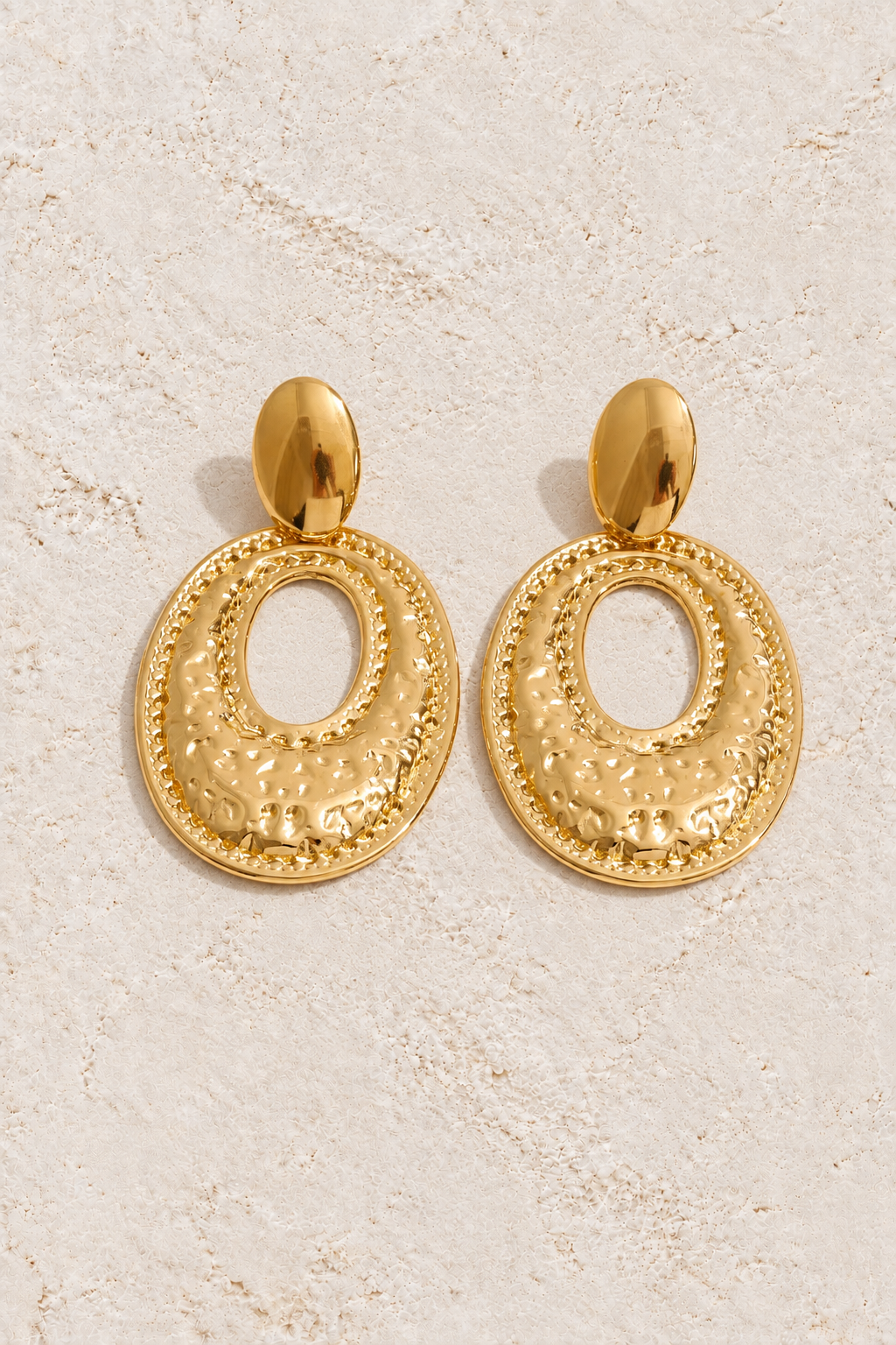 
                  
                    Gold hammered oval drop earrings on cream plaster
                  
                