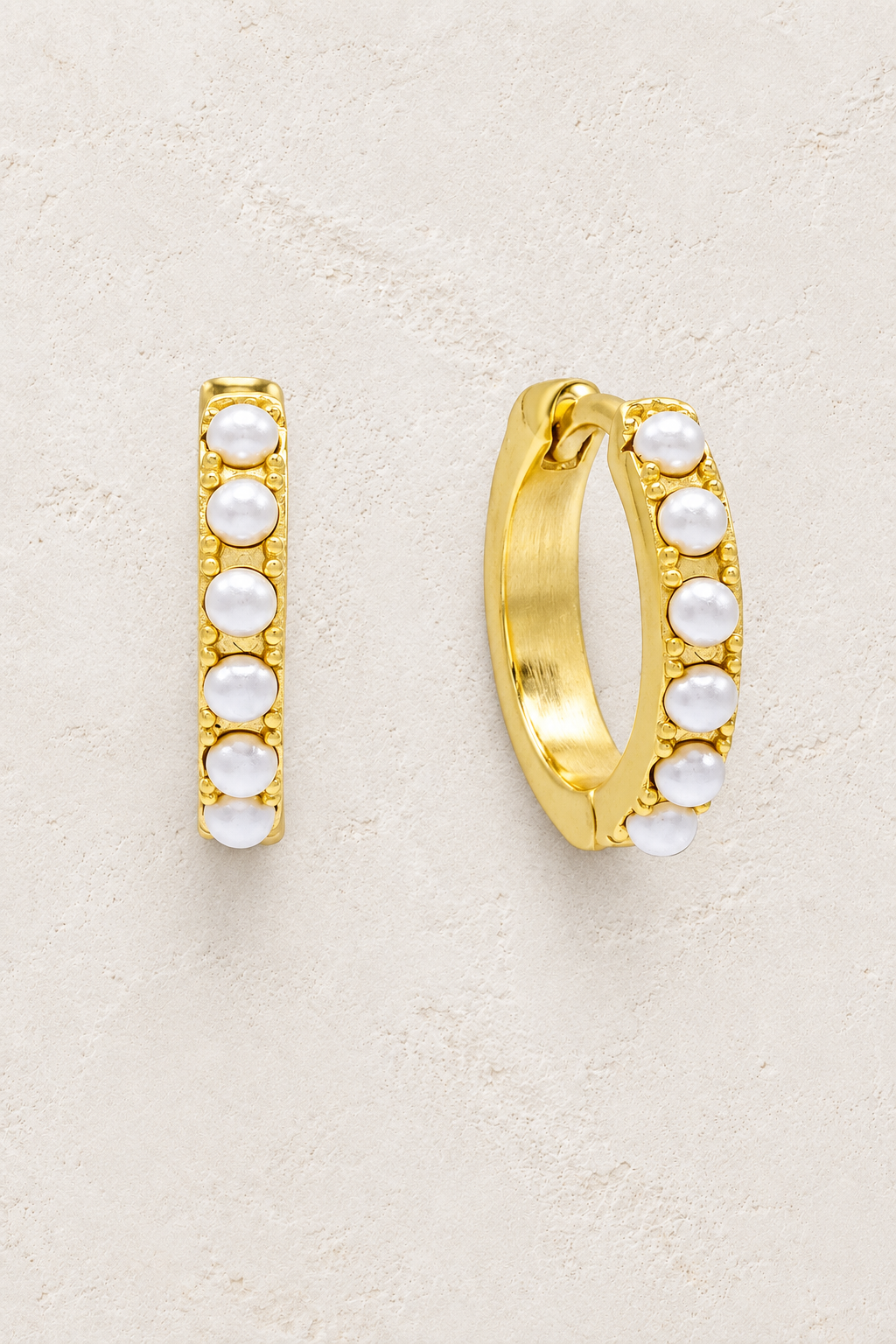 
                  
                    Gold plated sterling silver pearl huggie earrings on a warm off-white textured plaster background
                  
                