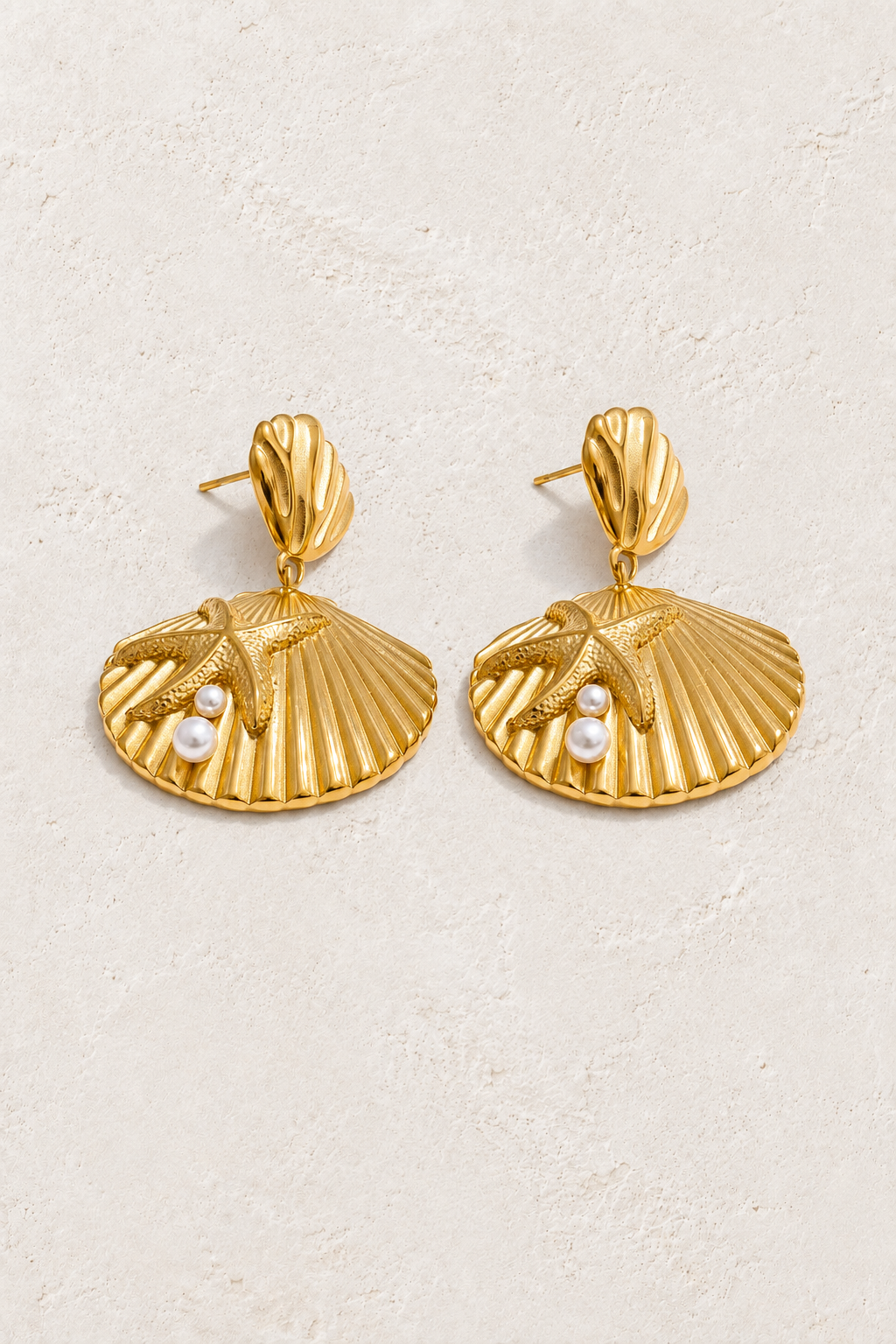 Gold shell fan and pearl stud earrings on a warm off-white textured plaster background