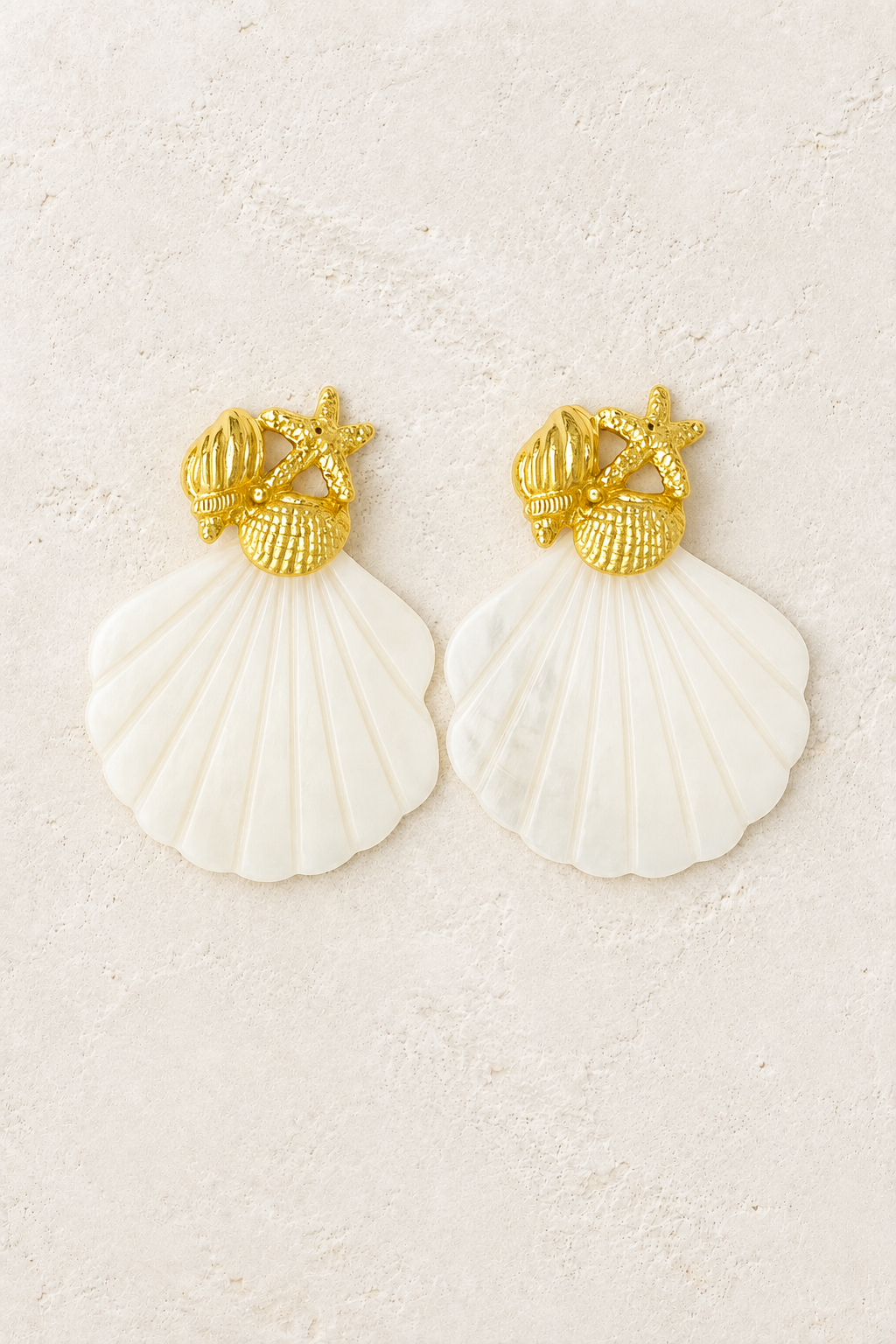 Gold starfish and pearly shell statement earrings on a warm off-white textured plaster background