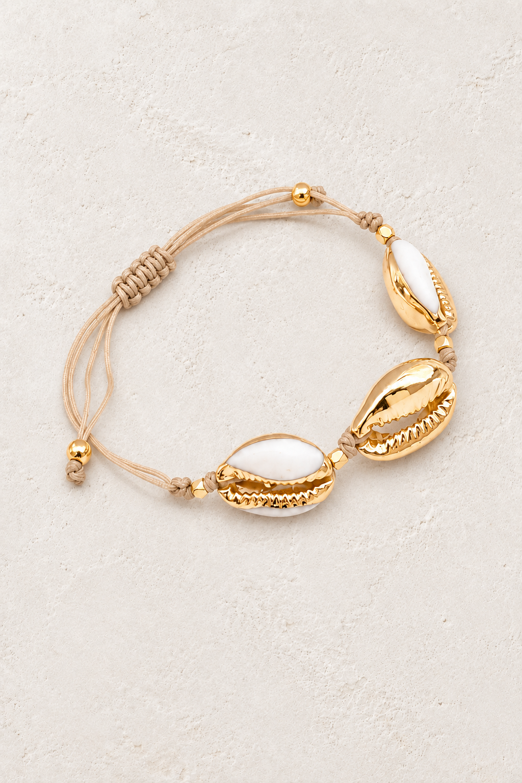
                  
                    Golden Tide gold cowrie shell cord bracelet on a warm off-white textured plaster background
                  
                