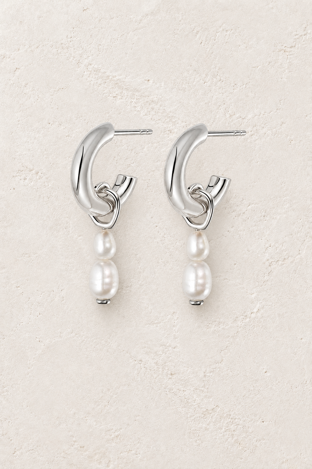 Sterling silver open hoop stud earrings with double pearl drops on a warm off-white textured plaster background
