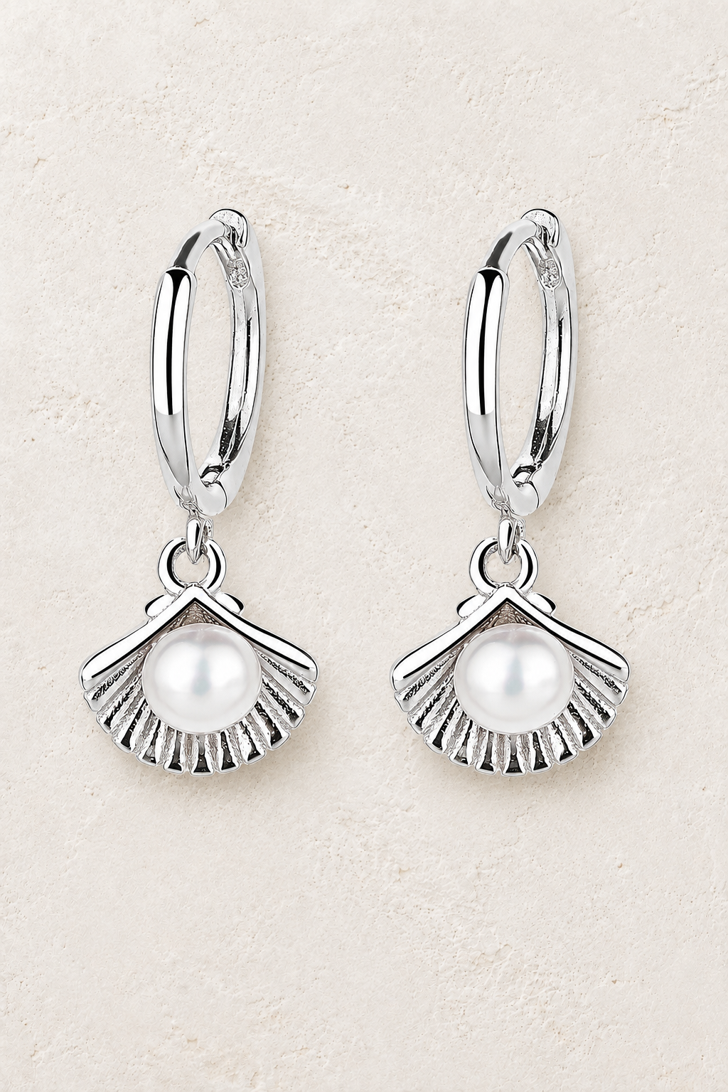 Sterling silver hoop earrings with fan shell and pearl charms on a warm off-white textured plaster background