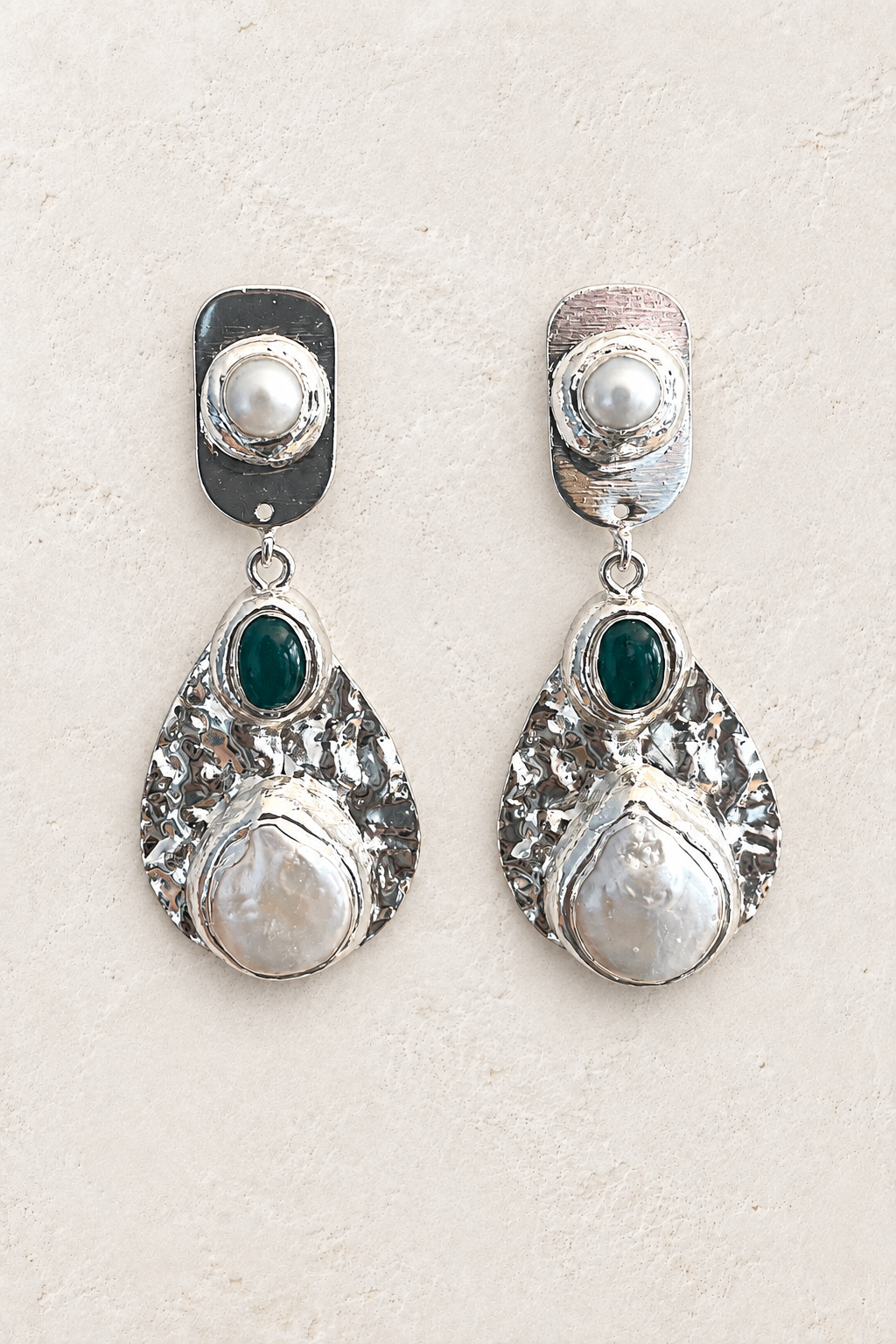 Silver hammered statement earrings with pearls and green stones on a warm off-white textured plaster background