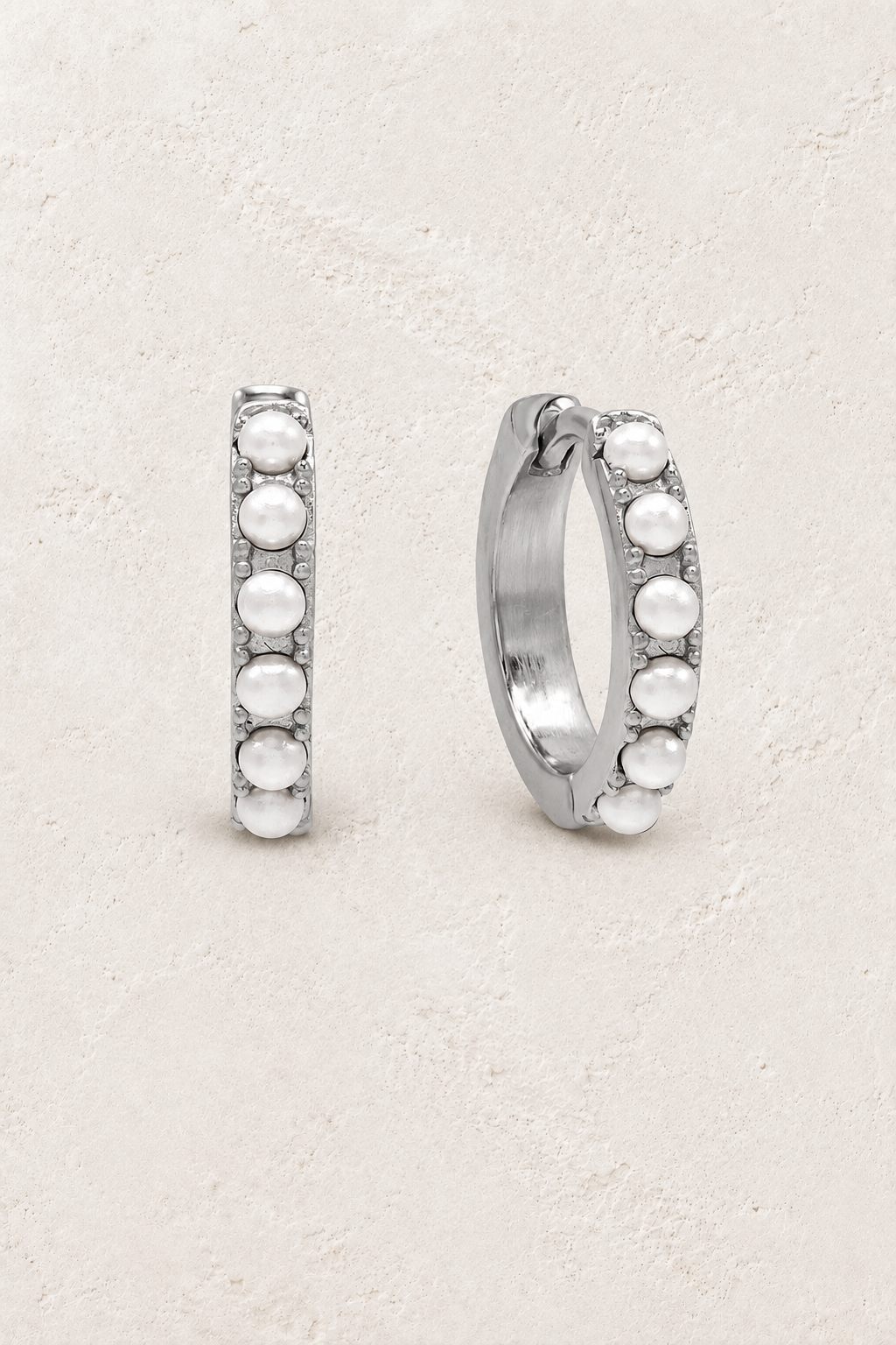 Sterling silver pearl huggie earrings on a warm off-white textured plaster background