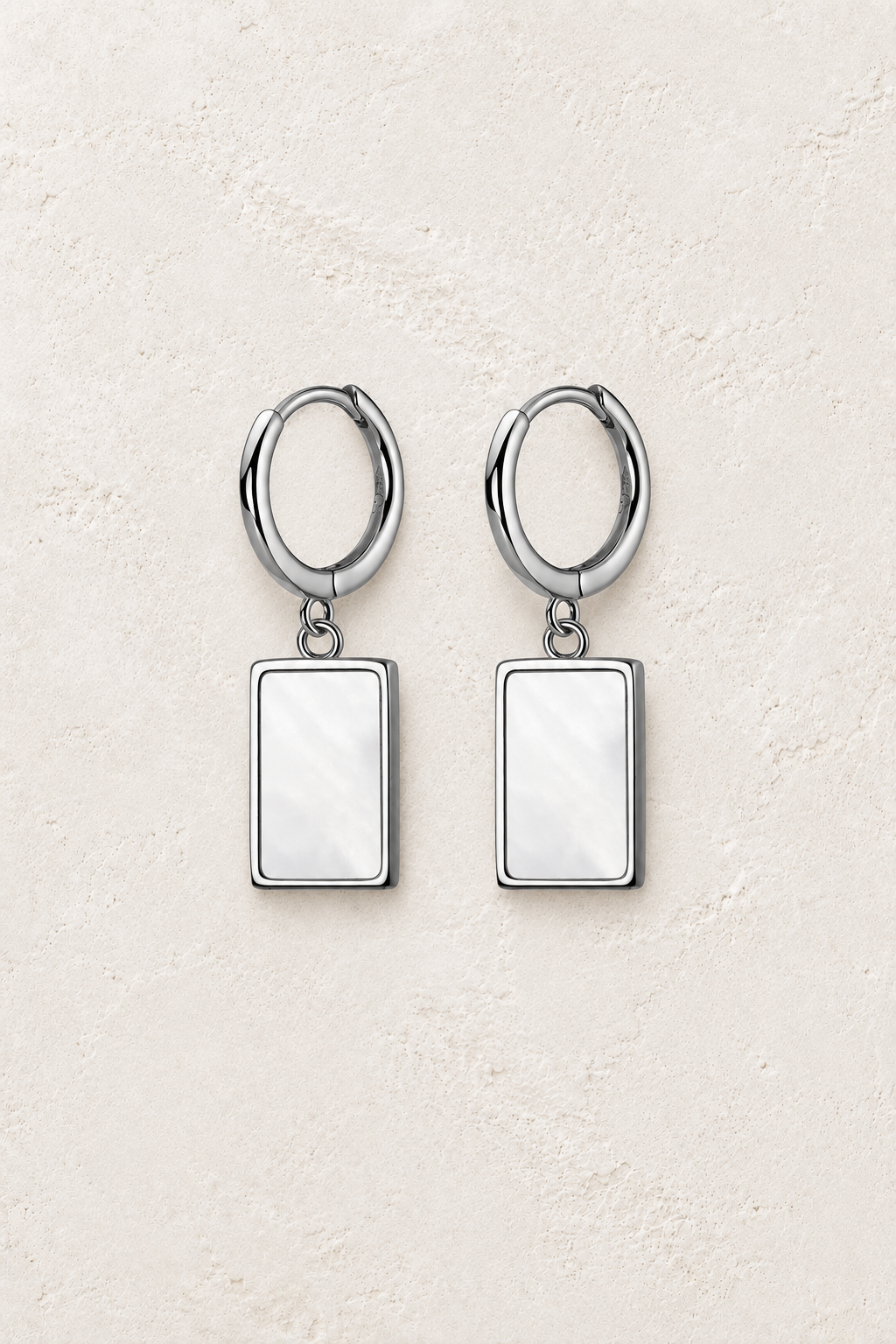Sterling silver huggie earrings with rectangular pearly shell drops on a warm off-white textured plaster background