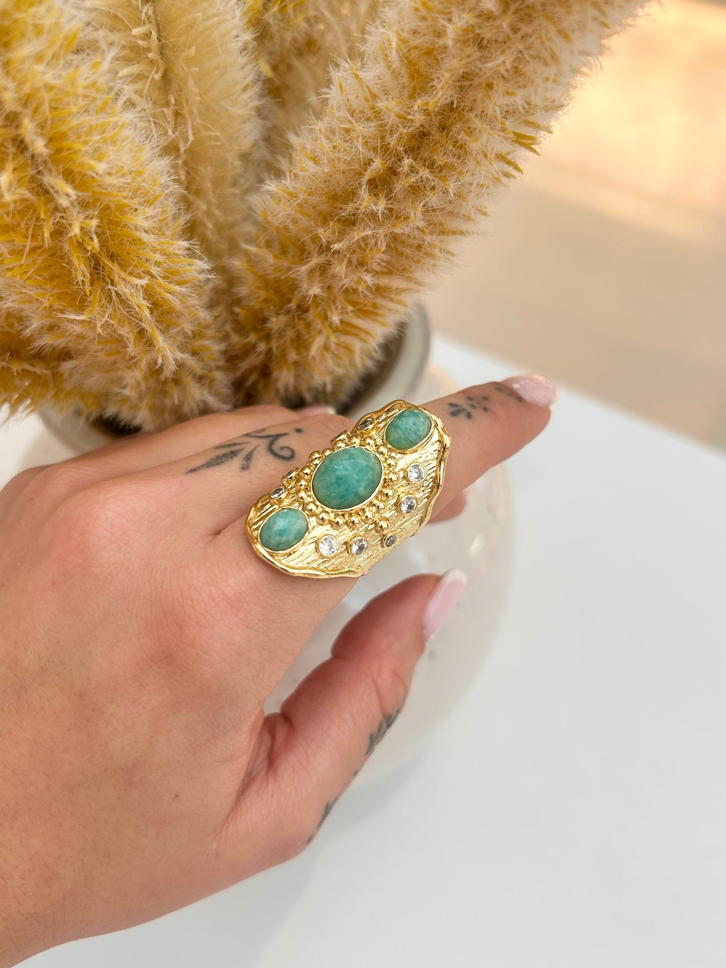 
                  
                    Hand wearing the Solara Amazonite Statement Ring featuring three natural aqua-green stones and crystal accents in a textured gold-plated design.
                  
                