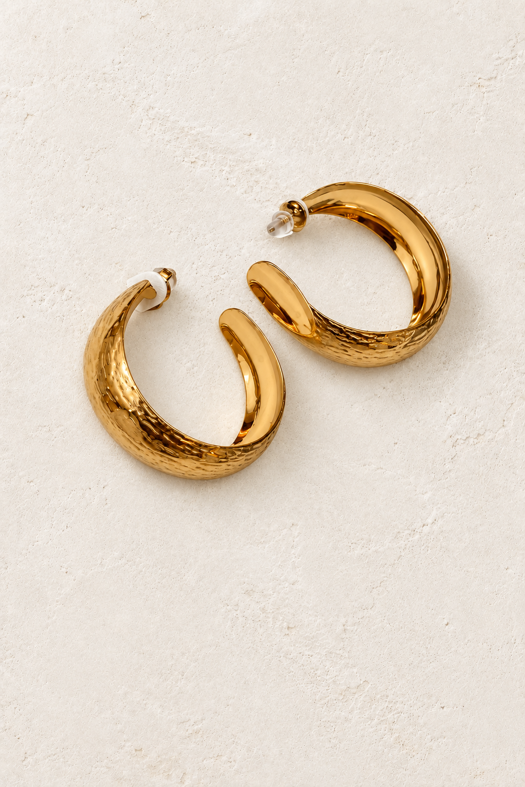 
                  
                    Zahara Hammered Gold Hoop Earrings on cream plaster background
                  
                