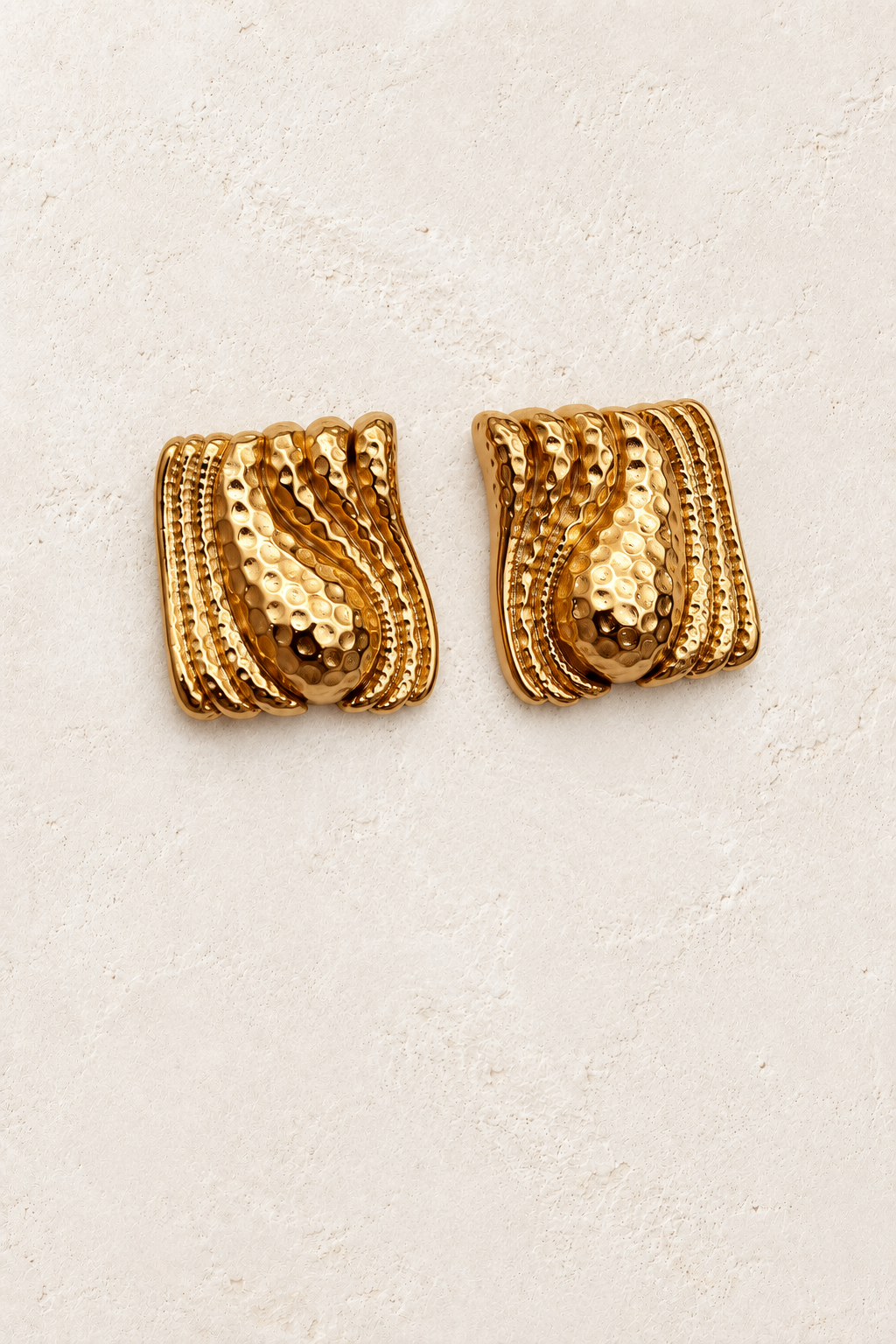 
                  
                    Zaria Hammered Gold Wave Stud Earrings on cream plaster background
                  
                