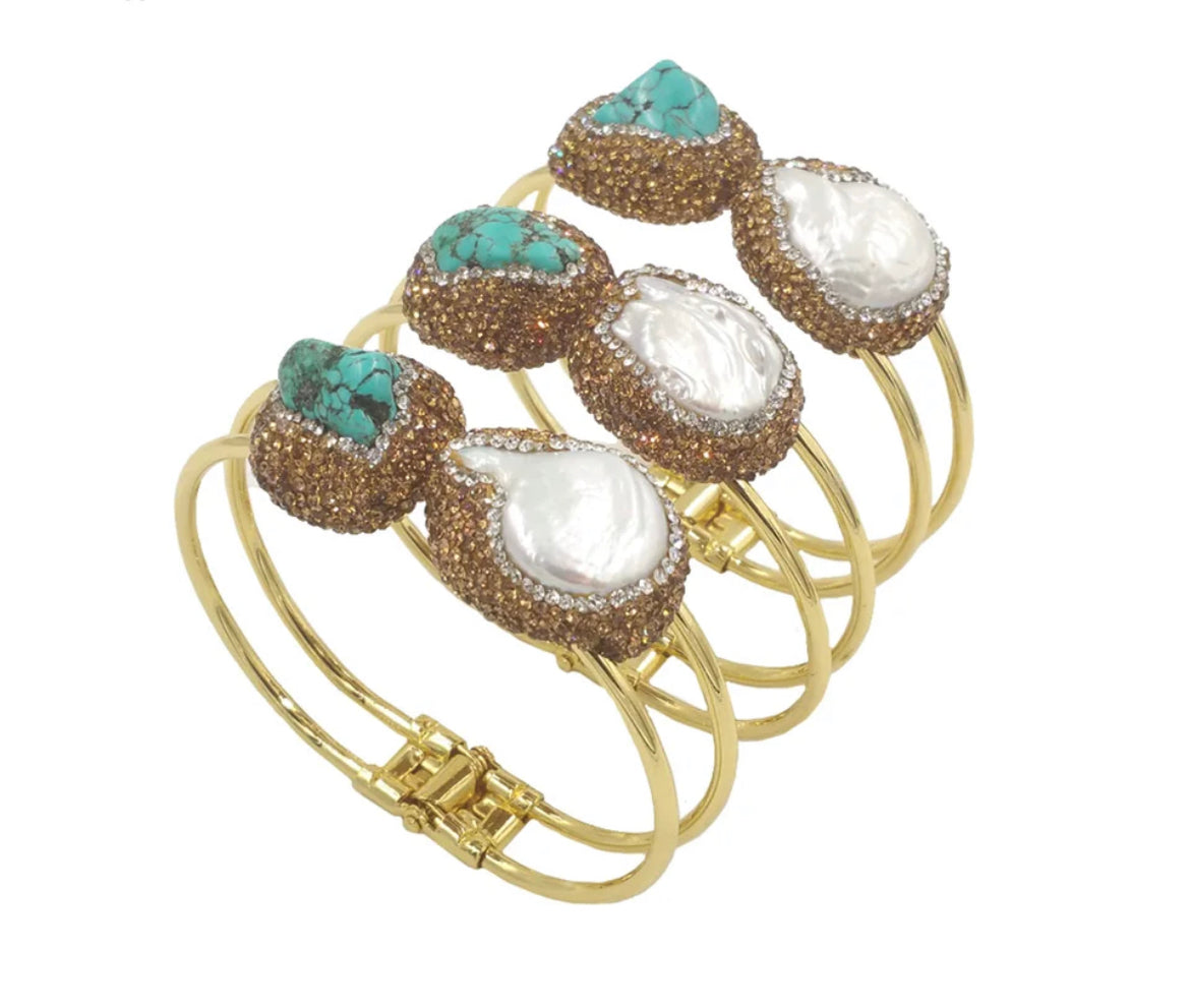 
                  
                    Multiple gold-plated bangle bracelet with two decorative oval charms, one featuring a white mother-of-pearl inlay and the other featuring a turquoise stone surrounded by pavé crystals, displayed on a white round pedestal.
                  
                
