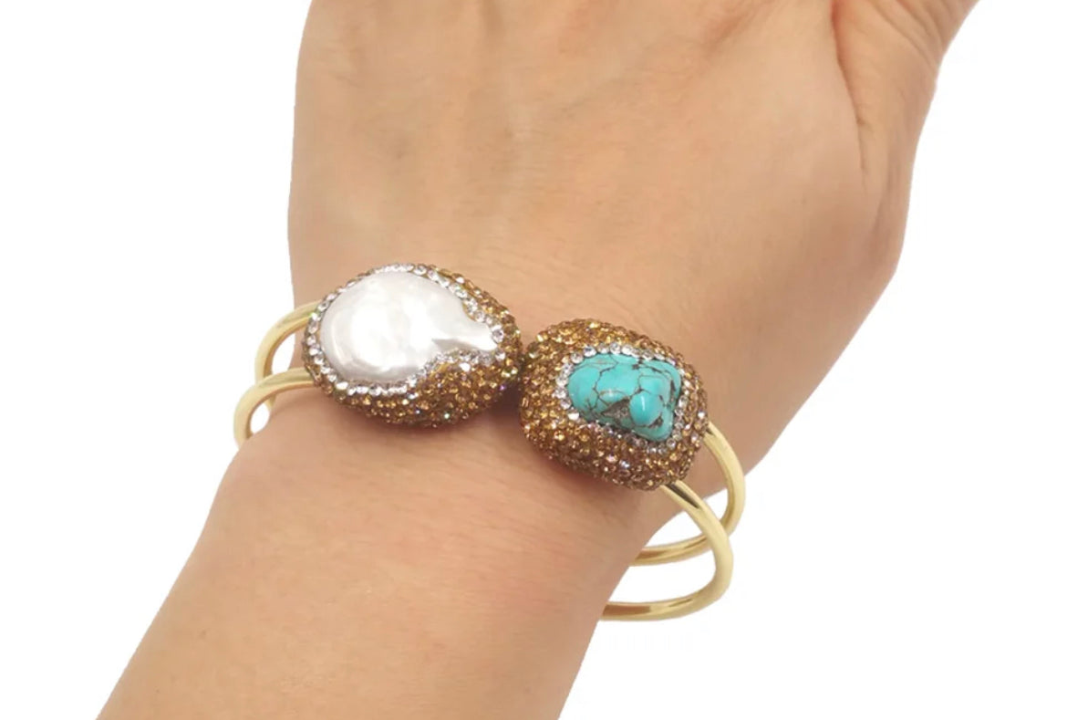 
                  
                    A gold-plated bangle bracelet with two decorative oval charms, one featuring a white mother-of-pearl inlay and the other featuring a turquoise stone surrounded by pavé crystals, displayed on a white round pedestal.
                  
                